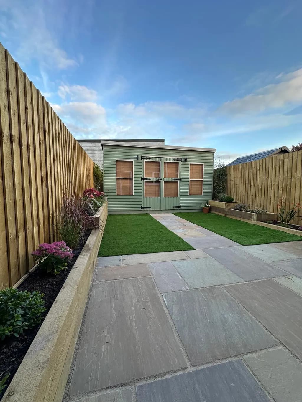 Artificial grass lawn bordered by tiered sleeper planters and a sandstone seating area and pathway.